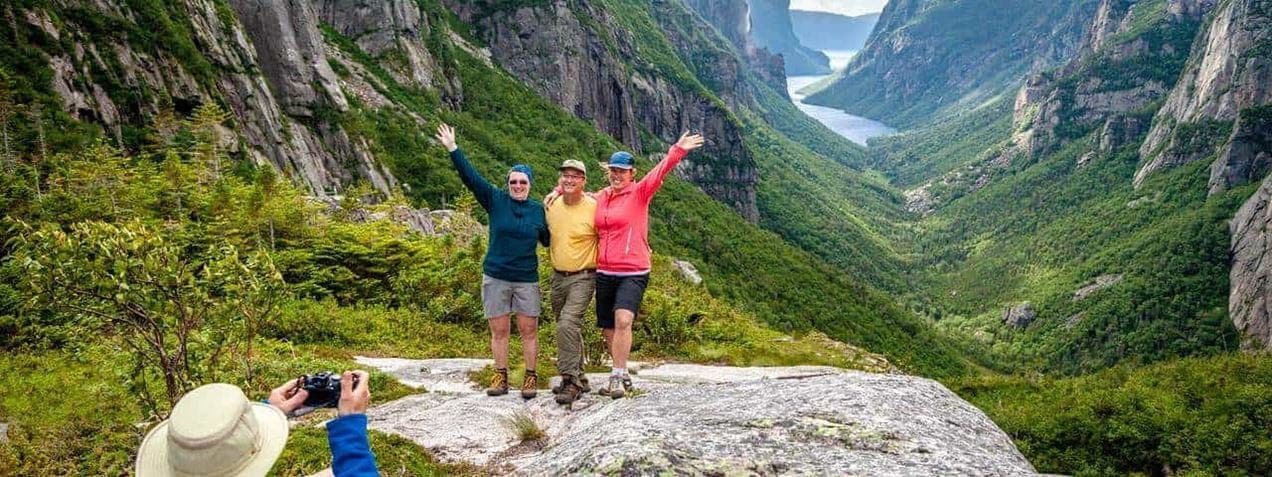 Three people pose on a rocky ledge with arms raised, surrounded by lush green mountains and a scenic valley. A person in the foreground takes their photo. A distant river winds through the landscape under a blue sky.